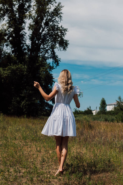 A girl in a rustic dress turned her back and holds flowers in her hand - Фото, изображение