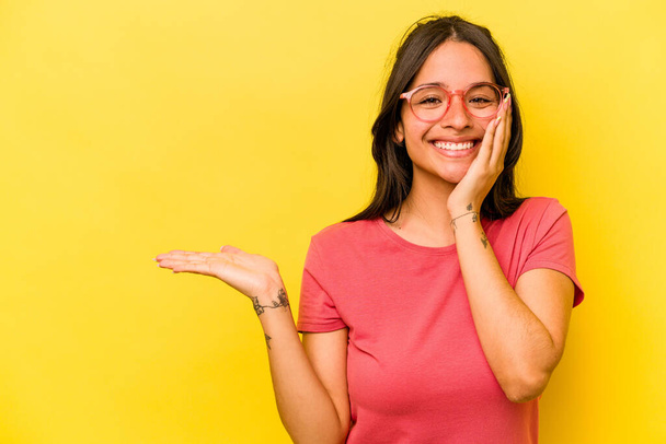 Young hispanic woman isolated on yellow background holds copy space on a palm, keep hand over cheek. Amazed and delighted. - Foto, immagini