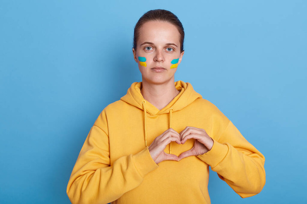 Attractive Caucasian woman wearing hoodie with blue and yellow flag on cheeks making heart shape gesture, expressing love to Ukraine, isolated over blue background. - Valokuva, kuva