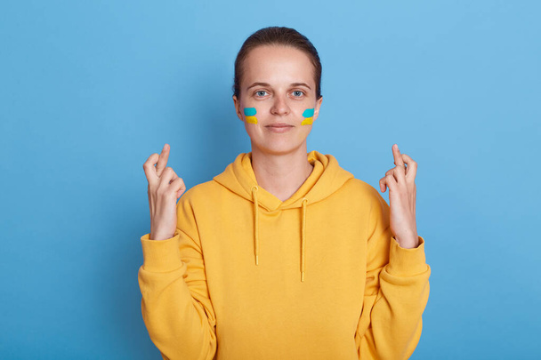 Adorable positive hopeful woman wearing hoodie with blue and yellow flag on cheeks, standing isolated over blue background and crossing fingers, making wish her country will free and win the war. - Foto, Imagem
