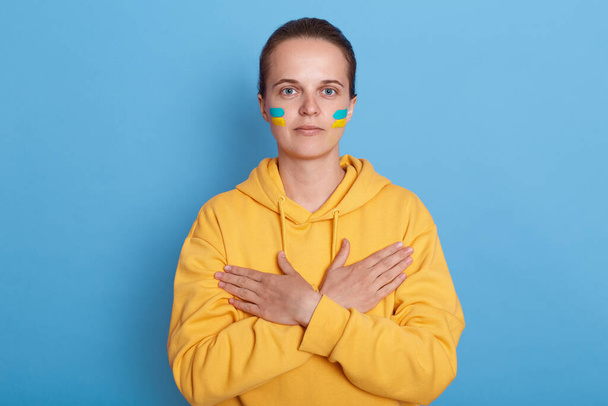 Portrait of serious woman wearing hoodie with blue and yellow flag on cheeks, isolated over blue background, keeps hands crossed on her chest, looking at camera. Glory to Ukraine. - Fotografie, Obrázek