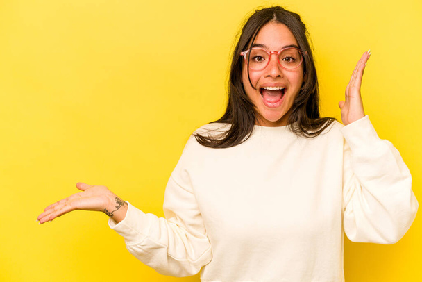 Young hispanic woman isolated on yellow background holds copy space on a palm, keep hand over cheek. Amazed and delighted. - Фото, изображение