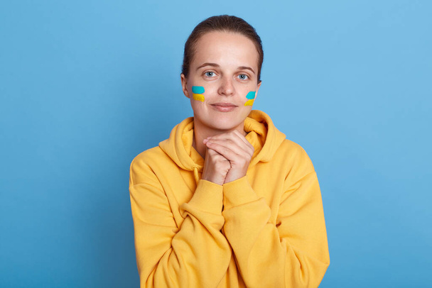Cute charming young adult woman wearing hoodie with blue and yellow flag on cheeks, standing isolated over blue background, keeps hands together under chin, support her native Ukraine. - Foto, Imagen