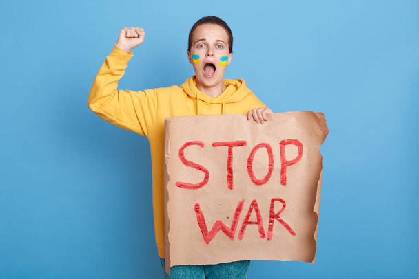 Excited patriotic woman in hoodie with Ukrainian flag on cheeks, posing isolated over blue background, protesting with placard with stop war inscription, screaming loud. - Photo, Image