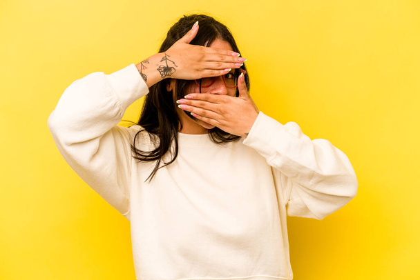 Young hispanic woman isolated on yellow background blink at the camera through fingers, embarrassed covering face. - Foto, imagen