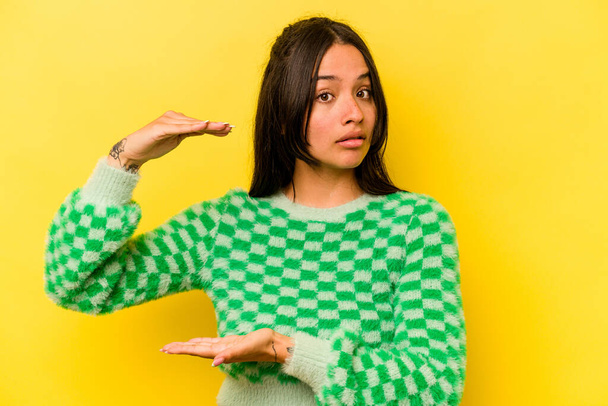 Young hispanic woman isolated on yellow background holding something with both hands, product presentation. - Fotografie, Obrázek
