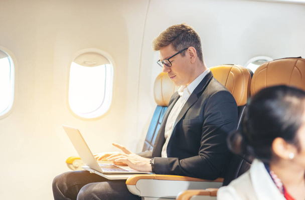 Tijdens een vlucht kijkt een zakenman in formele kledij en bril uit het raam terwijl hij aan een laptop werkt. Service met internettoegang aan boord en een business travel concept - Foto, afbeelding