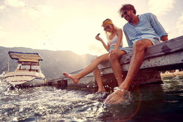 Playful couple sitting on wooden jetty by water, eating watermelon and making splashes in water.  Tourism, summertime, togetherness, lifestyle concept. - Foto, Imagem