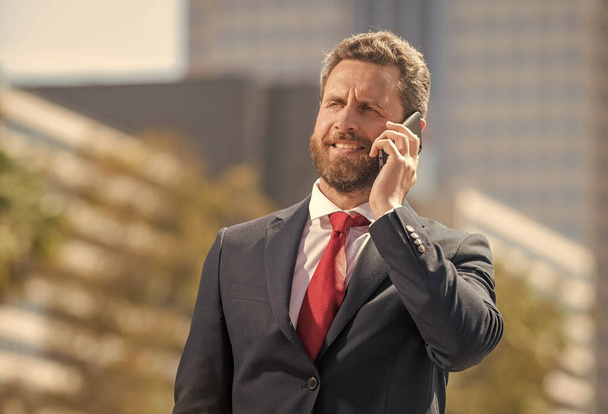 bebaarde zakenman in formele kleding heeft een telefoongesprek buiten kantoor, mobiele technologie - Foto, afbeelding