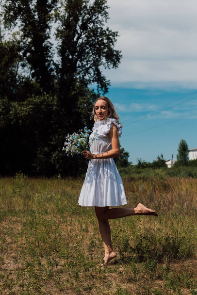 A young woman stands in a village, holding flowers in her hands and raises leg - Fotografie, Obrázek
