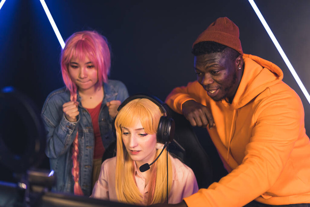 Three friends spending leisure time in a gaming club. Young blonde girl using professional headset and desktop computer to win a tournament online cheered up by her friends. - Valokuva, kuva