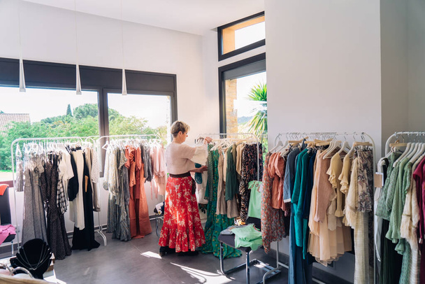 Dojrzała kobieta wybierająca ubrania z wieszaka, robiąca zakupy w sklepie z modą. koncepcja zakupów. - Zdjęcie, obraz