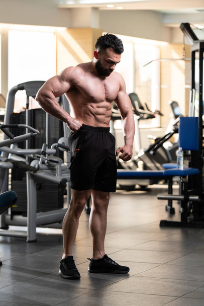 Handsome Young Man Standing Strong In The Gym And Flexing Muscles - Muscular Athletic Bodybuilder Fitness Model Posing After Exercises - 写真・画像