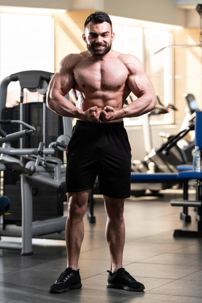Handsome Young Man Standing Strong In The Gym And Flexing Muscles - Muscular Athletic Bodybuilder Fitness Model Posing After Exercises - 写真・画像