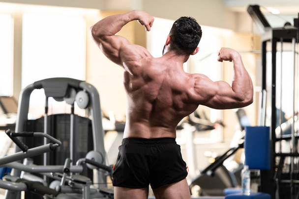Healthy Young Man Standing Strong In The Gym And Flexing Muscles - Muscular Athletic Bodybuilder Fitness Model Posing After Exercises - 写真・画像