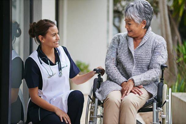 Langetermijnsteun voor de laatste jaren. Schot van een oudere vrouw in een rolstoel wordt voor door een verpleegster in een bejaardentehuis. - Foto, afbeelding
