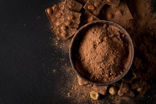 Broken chocolate nuts pieces and cocoa powder in bowl on dark stone background - Valokuva, kuva