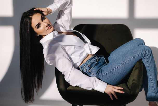 Female sitting on green chair. Her black hair is hanging down. Pretty woman in denim and white shirt - Фото, изображение