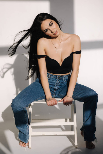 Sexy woman in black topic and blue jeans sits on chair during studio photoshoots. Brunette girl posing to photographer. Shadow on white background - Foto, afbeelding