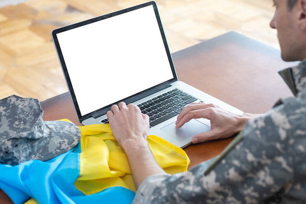 wojskowy człowiek gestykulujący w biurze przy laptopie z pustym ekranem, flaga Ukrainy. - Zdjęcie, obraz