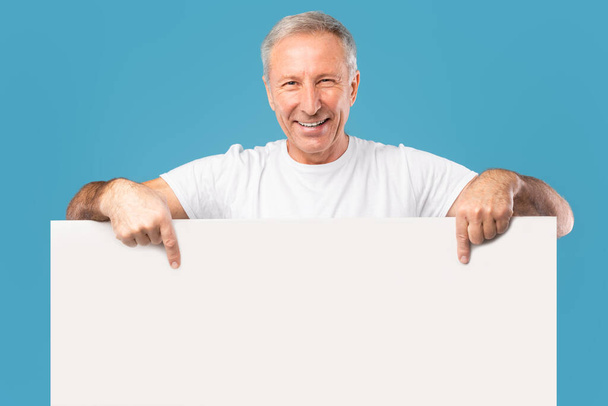 Ώριμος άντρας κρυφοκοιτάζει λευκή διαφημιστική πινακίδα στο στούντιο - Φωτογραφία, εικόνα