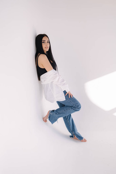 Pretty woman barefoot in studio. Beautiful brunette girl in black topic with white shirt and blue denim jeans. White background of cyclorama - Foto, Imagem