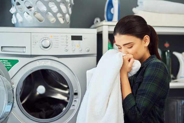 A friss szennyes illata. Vágott felvétel egy vonzó fiatal nőről, aki frissen mosott törölközőt szagolgat, miközben otthon mosogat.. - Fotó, kép