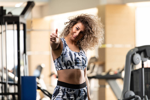 Attractive Young Woman Showing Thumbs Up While Standing Strong In The Gym And Flexing Muscles - Beautiful Athletic Fitness Model Posing After Exercises - Foto, Bild