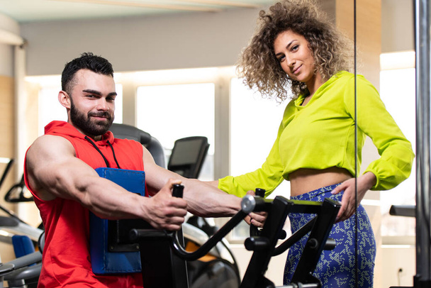 Young Man Working Out In Gym - Doing Back Exercise On Machine With Help Of Her Trainer - Zdjęcie, obraz
