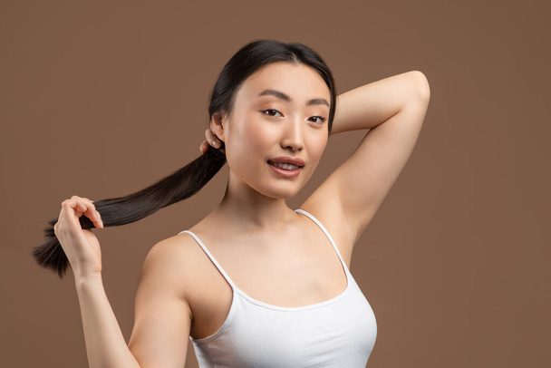 Jeune Coréenne à la peau parfaite montrant ses longs cheveux raides et soyeux, posant sur fond de studio marron - Photo, image
