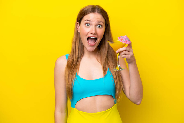 Young caucasian woman holding a cocktail isolated on yellow background with surprise and shocked facial expression - Fotografie, Obrázek