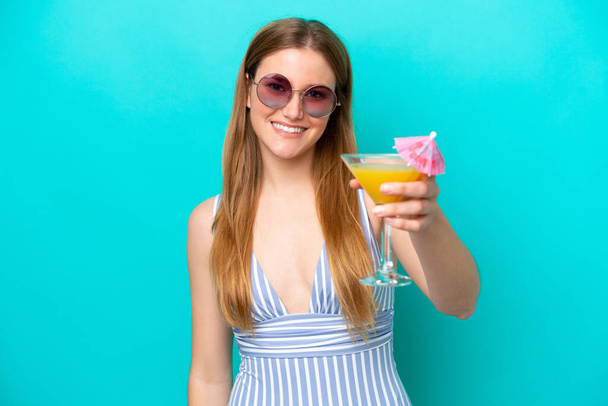 Young woman isolated on blue background in swimsuit and holding a cocktail - Photo, image