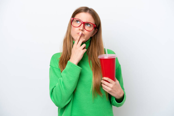 Молодая белая женщина, пьющая содовую на белом фоне, испытывает сомнения, глядя вверх. - Фото, изображение