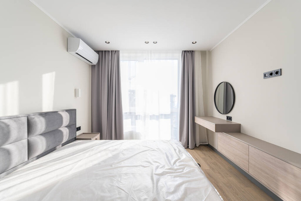 Amplo canto do quarto em estilo moderno, com uma cama grande, janela, ar condicionado e um espelho redondo - Foto, Imagem