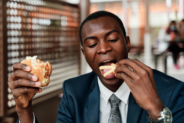 Happy young african american man having hamburger and french fries in fast food restaurant - Zdjęcie, obraz