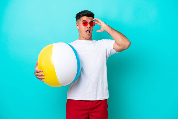 Молодий білий чоловік тримає пляжний м'яч ізольований на синьому фоні робить несподіваний жест, дивлячись в сторону
 - Фото, зображення