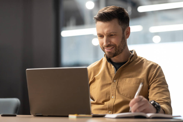 Χαρούμενος επιχειρηματίας χρησιμοποιώντας το laptop και κρατώντας σημειώσεις εργασίας στο χώρο εργασίας - Φωτογραφία, εικόνα