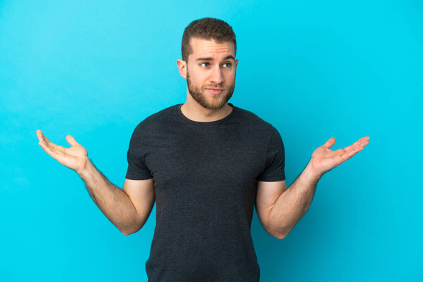 Young handsome caucasian man isolated on blue background having doubts while raising hands - Foto, immagini