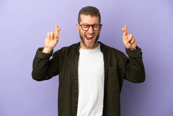 Young handsome caucasian man isolated on purple background with fingers crossing - Фото, изображение