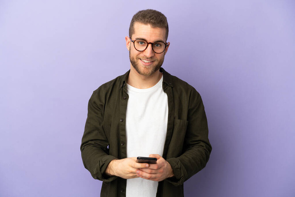 Молодой красивый белый мужчина, изолированный на фиолетовом фоне, отправляет сообщение с мобильным телефоном - Фото, изображение