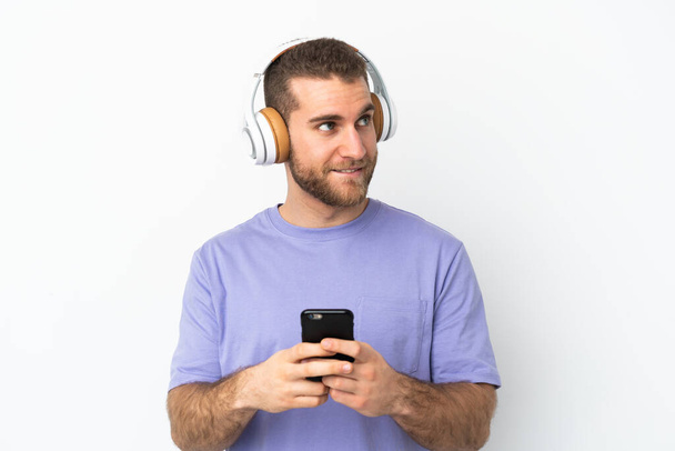 Young handsome caucasian man isolated on white background listening music with a mobile and thinking - Fotó, kép