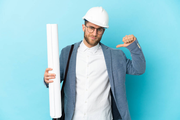 Молодий архітектор чоловік з шоломом і тримає креслення над ізольованим тлом, показуючи великий палець вниз з негативним виразом
 - Фото, зображення