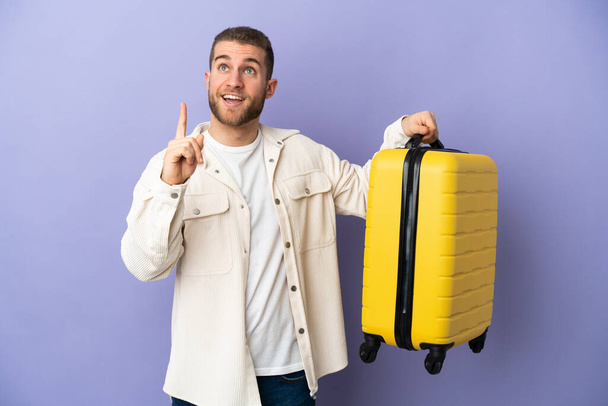 Молодий красивий кавказький чоловік ізольований на фіолетовому фоні у відпустці з подорожньою валізою і вказує
 - Фото, зображення