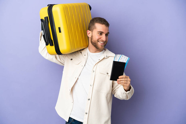 Junger schöner kaukasischer Mann isoliert auf lila Hintergrund im Urlaub mit Koffer und Pass - Foto, Bild