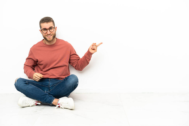 Mladý pohledný muž sedící na podlaze a ukazující prstem do strany - Fotografie, Obrázek