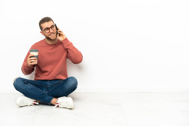 Joven hombre guapo sentado en el suelo sosteniendo café para llevar y un móvil - Foto, imagen