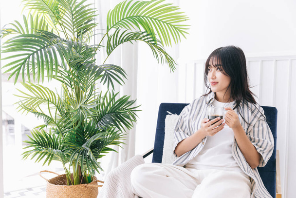 Young pretty woman smile and sits in the chair of the modern living area with a green plant, Relaxing drinking a cup of hot coffee and enjoying a quiet time and indoor environment at cozy home. - 写真・画像