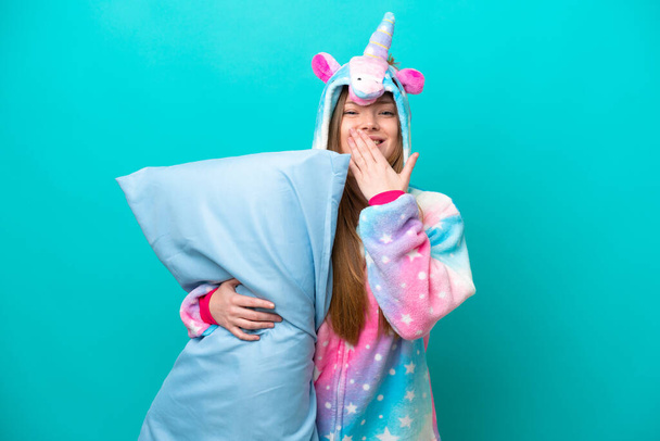 Young caucasian girl with unicorn pajamas holding pillow isolated on blue background happy and smiling covering mouth with hand - Photo, Image