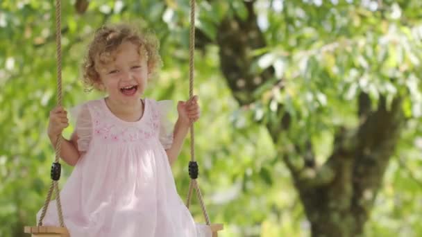 Spring and happy summer time. Joyful smiling little girl swinging on the swing, child with blue eyes and curly blond hair plays in the green garden at home, concept of healthy growth - Filmagem, Vídeo