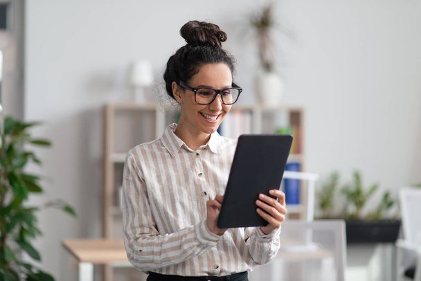 Позитивная молодая предпринимательница, пользующаяся цифровым планшетом в офисе и улыбающаяся, проверяя свои планы на блокноте - Фото, изображение